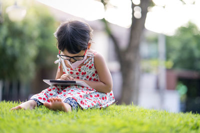 Little girl with protective face mask wearing wearing glasses sitting at field drawing on tablet.