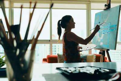 Side view of woman painting by window at home