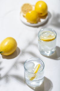 Two glass of water with ice cubes and lemon slices and a plate of lemons stand on a white table.