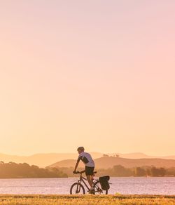 Man riding bicycle by lake against sky during sunset