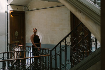 Pensive african american female standing in hall of shabby building with ornate railings and looking up