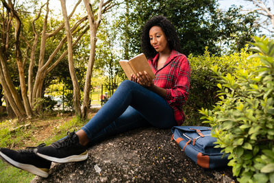 Young woman sitting on book by tree against plants