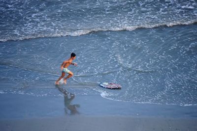 Side view of shirtless man running towards skimboard on shore at beach