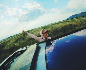 Cheerful young woman with arm raised leaning from car against sky