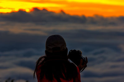 Rear view of woman with camera against cloudscape during sunset