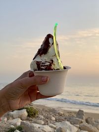 Hand holding ice cream cone at beach against sky