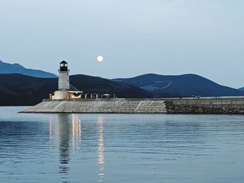 Lighthouse by river and buildings against clear sky