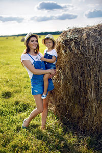 Daughter with mom standing in the field at the sheaf in hats and denim jumpsuits during sunset