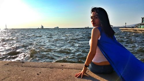 Portrait of woman sitting at harbor by sea against blue sky during sunny day