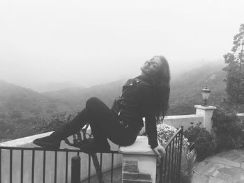 Woman sitting on railing by mountain against sky