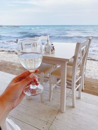 Hand holding glass of water at beach against sky