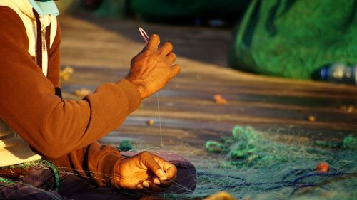 Midsection of fisherman sewing fishing net while sitting outdoors during sunny day