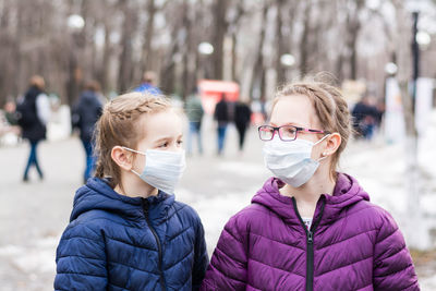 Two girls in protective face masks in the park on the background of a crowd of people