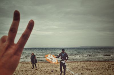 Cropped hand showing peace sign against man with kite at beach against cloudy sky