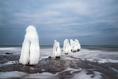 Frozen wooden posts in sea against cloudy sky during winter