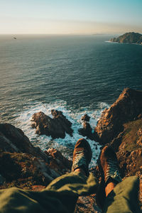 Low section of man sitting on mountain by sea against sky