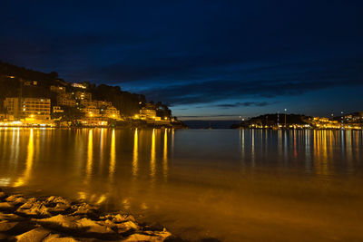 Illuminated buildings by sea against sky at night