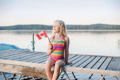 Happy caucasian girl sitting on pier by lake and waving canadian flag. kid celebrating canada day 
