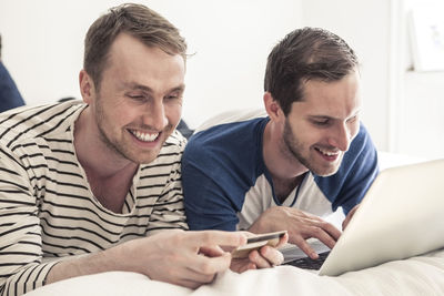 Happy gay men using credit card and laptop to shop online while lying in bed at home