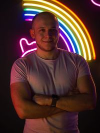 Portrait of smiling young man with arms crossed standing against illuminated light