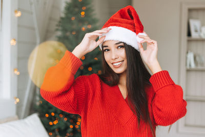 Young asian woman in cozy red knitted sweater and santa hat having fun in room with christmas tree