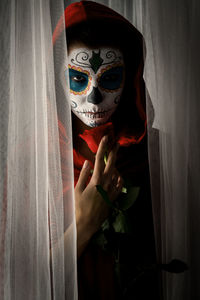 Portrait of woman with halloween make-up holding rose while standing amidst curtains