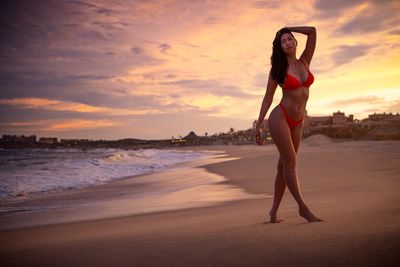Full length of woman wearing bikini standing at beach during sunset