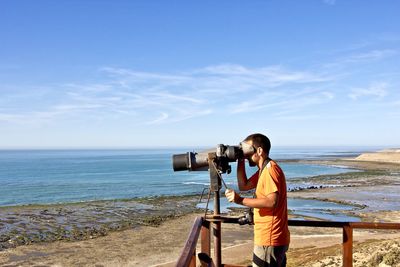 Side view of mature man using binoculars at beach against sky