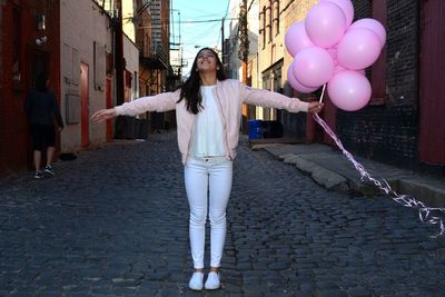 Happy woman holding pink helium balloons while standing on cobbled alley