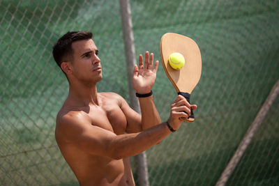 Shirtless young man playing tennis while standing against chainlink fence