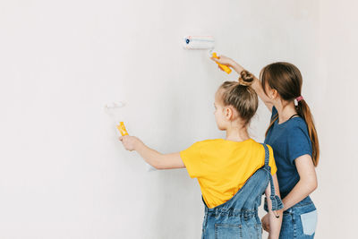 Two sisters are painting the walls of the house white, in denim overalls and a bright yellow t-shirt