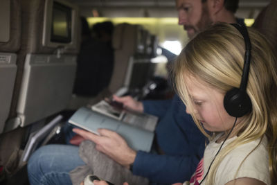 Side view of father reading magazine while daughter listening music in airplane