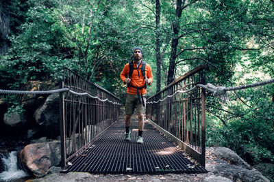 Carefree male tourist with backpack walking along metal suspension bridge and admiring amazing nature of woods during vacation