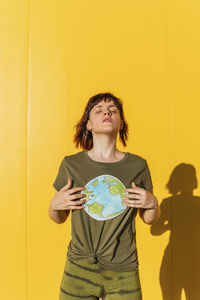 Mid adult woman with eyes closed holding planet earth drawing in front of wall during sunny day