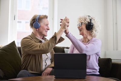 Senior man and woman giving each other high five while sitting in living room in front of digital tablet