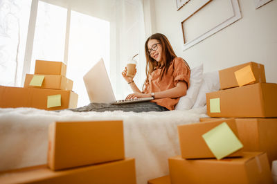 Low angle view of young woman with coffee using laptop on bed in home office