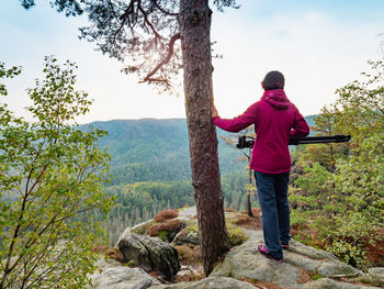 Photographer woman in red jacket stay with camera on tripod on cliff and thinking. woman hold tree