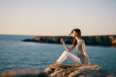 Woman sitting on rock by sea against sky during sunset