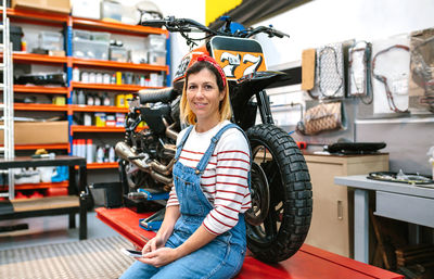 Mechanic woman holding phone sitting over platform with motorcycle on factory