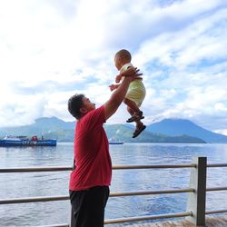 Side view of father holding baby mid-air against lake