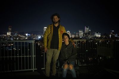 Portrait of man standing by woman sitting on chair against sky at night