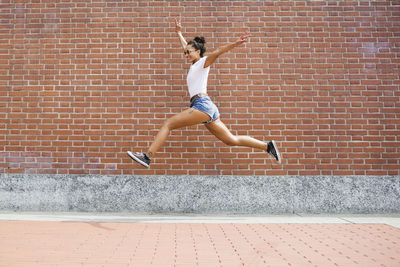 Exuberant young woman jumping in front of a brick wall