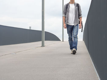 Low section of man walking on footbridge against sky