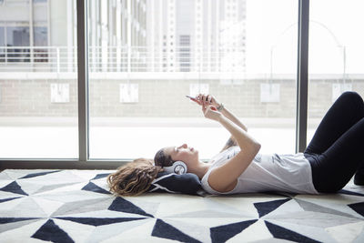 Side view of young woman using smart phone while lying on carpet by window at home
