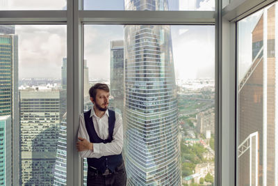 Thoughtful businessman with beard stands next to  window in office backdrop of skyscrapers in moscow