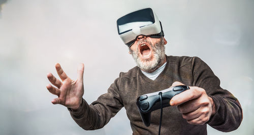 Senior man shouting while playing game with virtual reality headset against white background