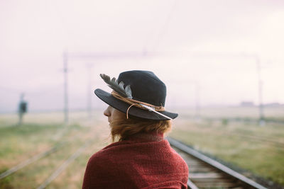 Rear view of woman wearing hat standing on railroad track against sky