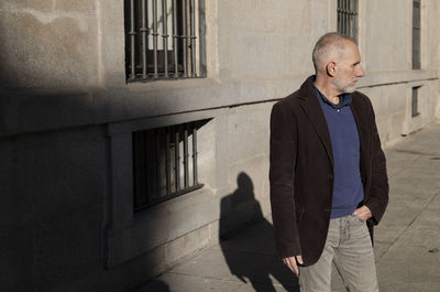 Adult man in suit standing on street against wall with sunlight and shadow