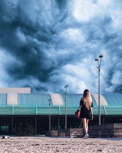 Rear view of woman walking on street against cloudy sky