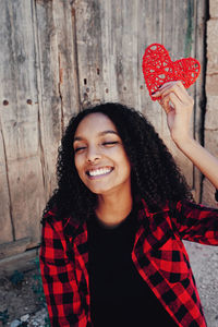 Cheerful woman holding red heart shape against wooden wall